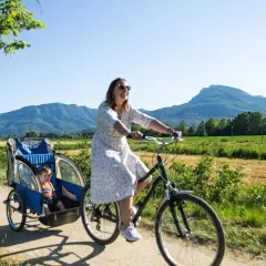 Disfrutando de un día soleado en la ruta el carrilet. Una mujer pedalea con un niño en un remolque, mientras otros ciclistas y un corredor disfrutan del paisaje montañoso.