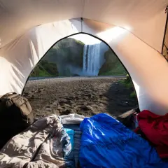 Vista desde una tienda de campaña con sacos de dormir y mochilas, con una cascada imponente al amanecer. ¡Una experiencia de acampada libre Europa inolvidable!