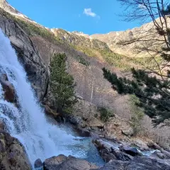 Cascada imponente en un paisaje montañoso, ideal para explorar rutas vielha. El agua cae sobre rocas y vegetación, con árboles y picos nevados al fondo.