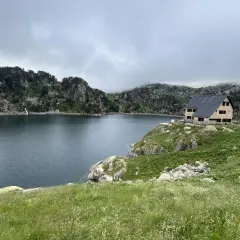 Refugio de montaña junto a un lago rodeado de montañas rocosas y vegetación.