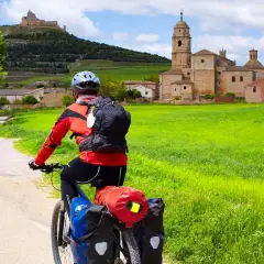 Ciclista en el Camino de Santiago en bici, con un pueblo y un castillo al fondo, rodeado de campos verdes.