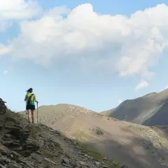 Senderista en la ruta Vall de Núria, con mochila y bastones, ascendiendo por un sendero rocoso con montañas al fondo.
