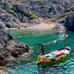 Barca amarilla en cala de aguas turquesas en el **Cabo de Creus**. Rocas escarpadas y gente en la playa al fondo.
