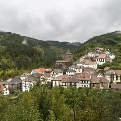 Pueblo pintoresco en el Pirineo Navarro, con casas de piedra y tejados rojos, rodeado de bosques frondosos y montañas nubladas.
