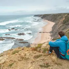 Persona con mochila azul contempla la costa salvaje durante un senderismo Portugal. Olas rompen en la playa desierta.