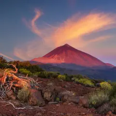 Volcán Teide bañado por el atardecer, ideal para actividades de montaña. Nubes anaranjadas y vegetación árida en primer plano.