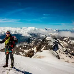 Alpinista en la cima del Gran Paradiso, con piolet y equipo, disfrutando de vistas espectaculares de montañas nevadas y nubes.