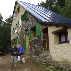 Llegada al refugio de Vallferrera. Dos jóvenes con mochilas y un hombre sentado revisando un mapa esperan en la entrada de piedra y estuco.