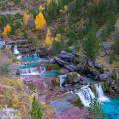 Cascada en las gradas de soaso, con aguas turquesas y rocas rojizas, rodeada de árboles otoñales y pinos verdes.