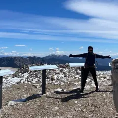 Persona amb els braços oberts al cim del Pedra dels Quatre Batlles, amb paisatge muntanyós i cel blau.