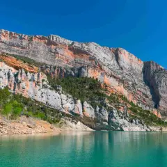 Majestuosas paredes rocosas de la sierra del Montsec se reflejan en las aguas turquesas de un embalse.
