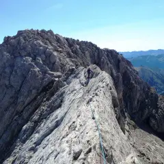 Alpinista en la cresta rocosa del Pico de Vallibierna, con un arnés y cuerda azul. Paisaje montañoso al fondo.