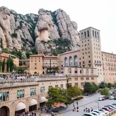 Edificios del monasterio se alzan bajo las imponentes rocas del parc natural de la muntanya de Montserrat. Turistas pasean por la plaza.
