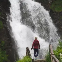 Mujer contemplando una cascada en Vielha e Mijaran. El agua cae con fuerza sobre rocas oscuras, rodeada de vegetación verde.