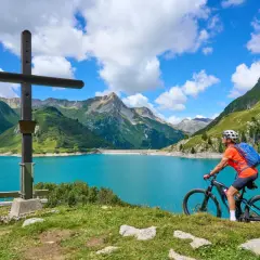 Ciclista en el Alpe Adria Radweg, junto a un lago turquesa y montañas verdes.