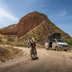 Ciclista pedaleando por un camino de badlands gravel, con una furgoneta camper aparcada al fondo.