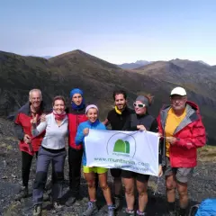 Grupo de senderistas celebra en la cima de una montaña, con el valle de **Aran** de fondo. Sostienen una pancarta de Muntania.com.