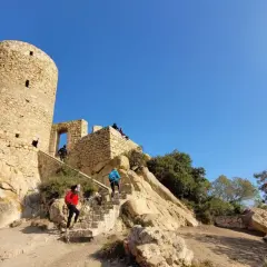 Personas suben escaleras hacia el histórico castillo de Burriac, una torre de piedra antigua bajo un cielo azul claro.