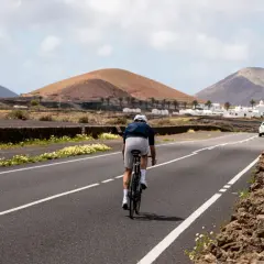 Ciclista pedaleando por una carretera en Lanzarote, con volcanes y casas blancas al fondo. Una aventura de lanzarote en bici.