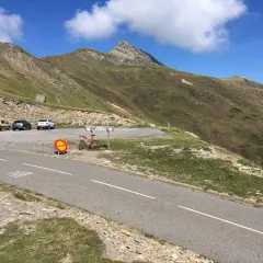 Un ciclista descansa junto a su bicicleta roja en el **puerto de Larrau**, con coches aparcados y montañas verdes bajo un cielo azul.