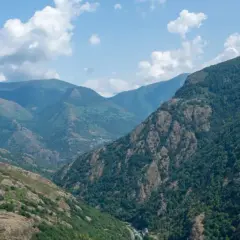 Paisaje montañoso con el bosque de Carlac cubriendo las laderas. Nubes blancas flotan en un cielo azul claro.