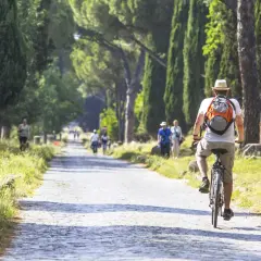 Un hombre pedalea por un camino empedrado en Roma, rodeado de altos cipreses y ruinas antiguas.