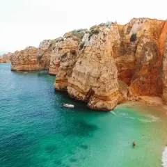 Aguas turquesas y acantilados dorados en las **playas Algarve**. Una pequeña barca navega cerca de una cala escondida donde una persona nada.
