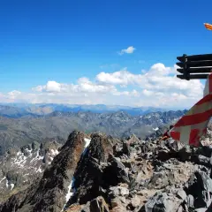 Cruz metálica en la cima de una pica d'estats, con bandera ondeando y un paisaje montañoso al fondo.