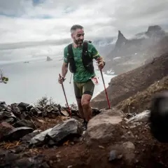Corredor de montaña con bastones asciende un sendero rocoso, superando un gran desnivel positivo con el mar y acantilados al fondo.