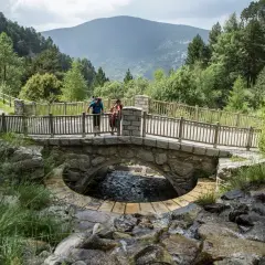 Pareja en un puente de piedra sobre un arroyo en Les Pardines, Andorra. Montañas y bosques al fondo.