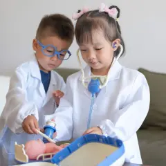Dos niños, vestidos de doctores, atienden a un muñeco. La niña usa un estetoscopio, mientras el niño le administra una inyección. Es una escena de juego de roles, una brecha de Rolando con niños.