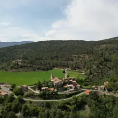 Vista aérea del pintoresco pueblo de **Vall de la Vansa**, rodeado de verdes prados y frondosos bosques, con casas de piedra y una iglesia.