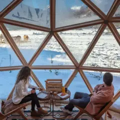 Pareja disfruta de un paisaje nevado en el Pirineo francés desde una cúpula geodésica. Vacas pastan en la distancia.