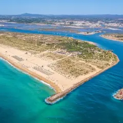 Vista aérea de la **isla de Tavira**, con su extensa playa de arena dorada, aguas turquesas y el canal que la separa de tierra firme, donde se aprecian salinas y la ciudad al fondo.