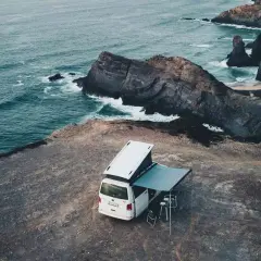 Furgoneta blanca con toldo y mesas en acantilado, disfrutando de la acampada libre Portugal con vistas al mar y rocas.