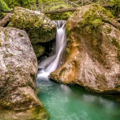 Cascada en las gargantas de Kakueta, con agua turquesa y rocas cubiertas de musgo. Un puente metálico cruza sobre la caída de agua.