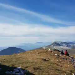 Dos personas contemplan el paisaje montañoso desde el Puig d'Estela, con un muro de piedra a la izquierda y el cielo azul con nubes.
