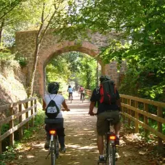 Ciclistas disfrutan de la vía verde Olot Girona, pedaleando bajo un antiguo arco de piedra rodeado de exuberante vegetación.