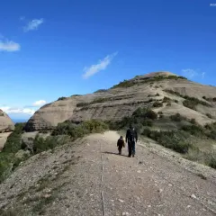 Un padre y su hijo caminan por un sendero rocoso hacia la cima del Montcau, bajo un cielo azul con nubes dispersas.