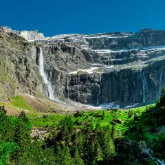 Cascadas impresionantes caen por acantilados rocosos cerca del pueblo de Gavarnie. Un sendero serpentea entre árboles verdes y prados exuberantes.