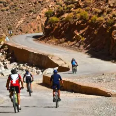 Ciclistas disfrutan de una aventura de **Marruecos en bici** por un camino sinuoso entre montañas rocosas y vegetación escasa.