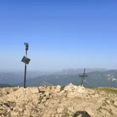 En el cap de la gallina pelada, un poste con placa y un sol solar vigilan el paisaje montañoso. A su lado, una cruz metálica se alza sobre rocas.