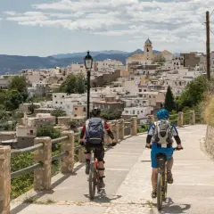 Dos ciclistas disfrutan de una ruta cicloturista ascendiendo hacia un pueblo blanco en la ladera de una montaña.