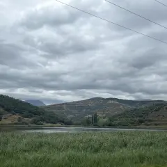 Paisaje sereno con un lago rodeado de colinas verdes y secas, bajo un cielo nublado. Las montañas a lo lejos parecen la geganta adormida.