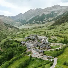 Pueblo de casas de piedra en un valle verde del Val d'Aran, rodeado de montañas cubiertas de vegetación y algunas cumbres nevadas.