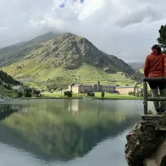Persona contempla el sereno lago en el valle de Nuria, con imponentes montañas verdes y un complejo de edificios al fondo.