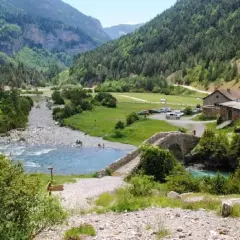 Un río de aguas turquesas fluye por un valle verde, cruzado por un puente de piedra. La ruta Bujaruelo invita a disfrutar de este paisaje montañoso.