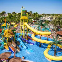 Divertido camping parque acuático con toboganes amarillos y azules, palmeras y gente disfrutando del sol y el agua.