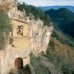 Ermita en la costa pubilla, aferrada a un acantilado rocoso con vistas a un valle boscoso. Una campana cuelga sobre la entrada.
