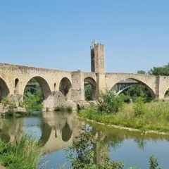 Puente medieval de Besalú, parte de una ruta histórica. Reflejo en el río y vegetación exuberante.